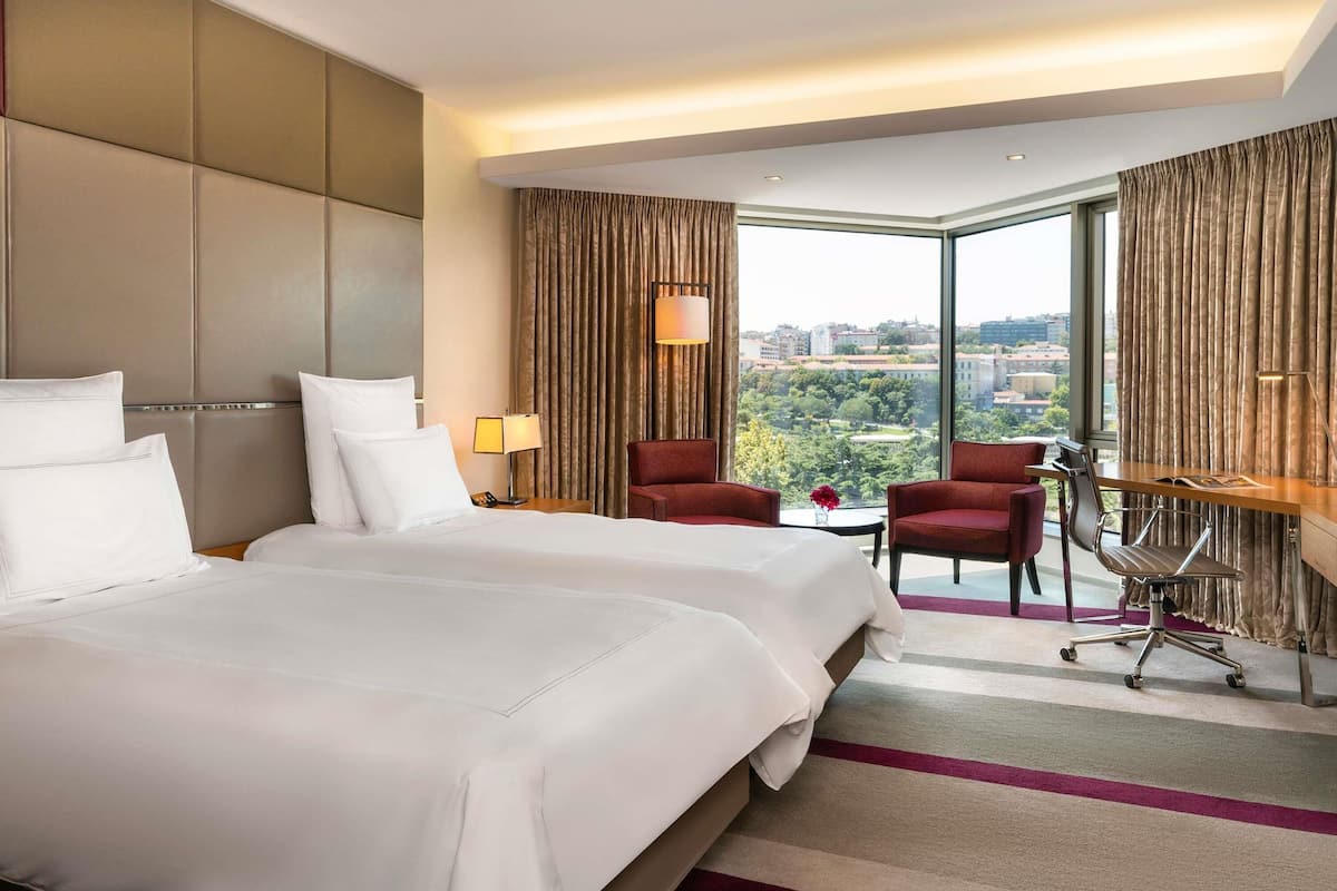 A hotel room with two beds, a desk, two chairs, a lamp, and a view of the city.