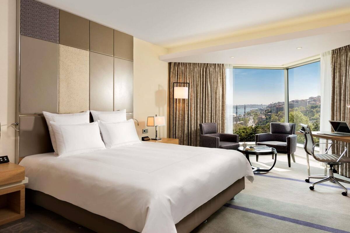 A hotel room with a large bed, a desk with a laptop, a chair, and a view of a cityscape.