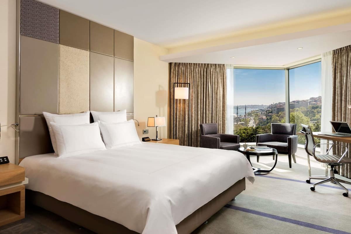 A hotel room with a large bed, a desk with a laptop, a chair, and a view of a cityscape.