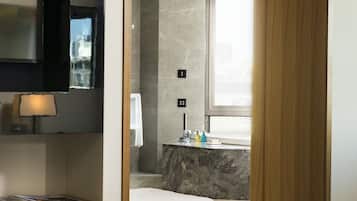 A modern hotel room with a large bed, a wooden-framed mirror, a bedside table with a lamp, and a view of the bathroom with a shower and a sink.