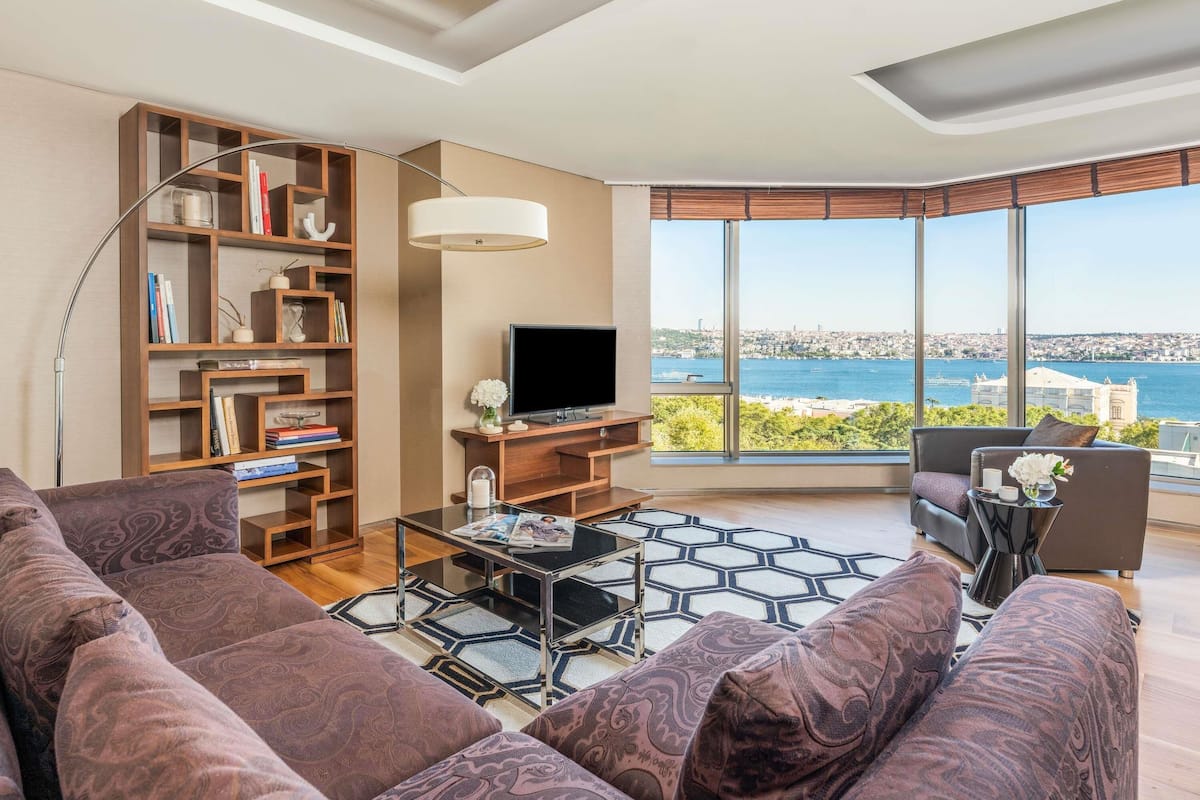 A modern living room with a large window offering a city view, a bookshelf, a flat-screen TV, and a sofa.