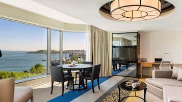 Una habitación de hotel moderna con un ventanal grande que ofrece vistas a la ciudad, un área de comedor con una mesa y sillas, un sofá, una mesa de centro y un televisor.