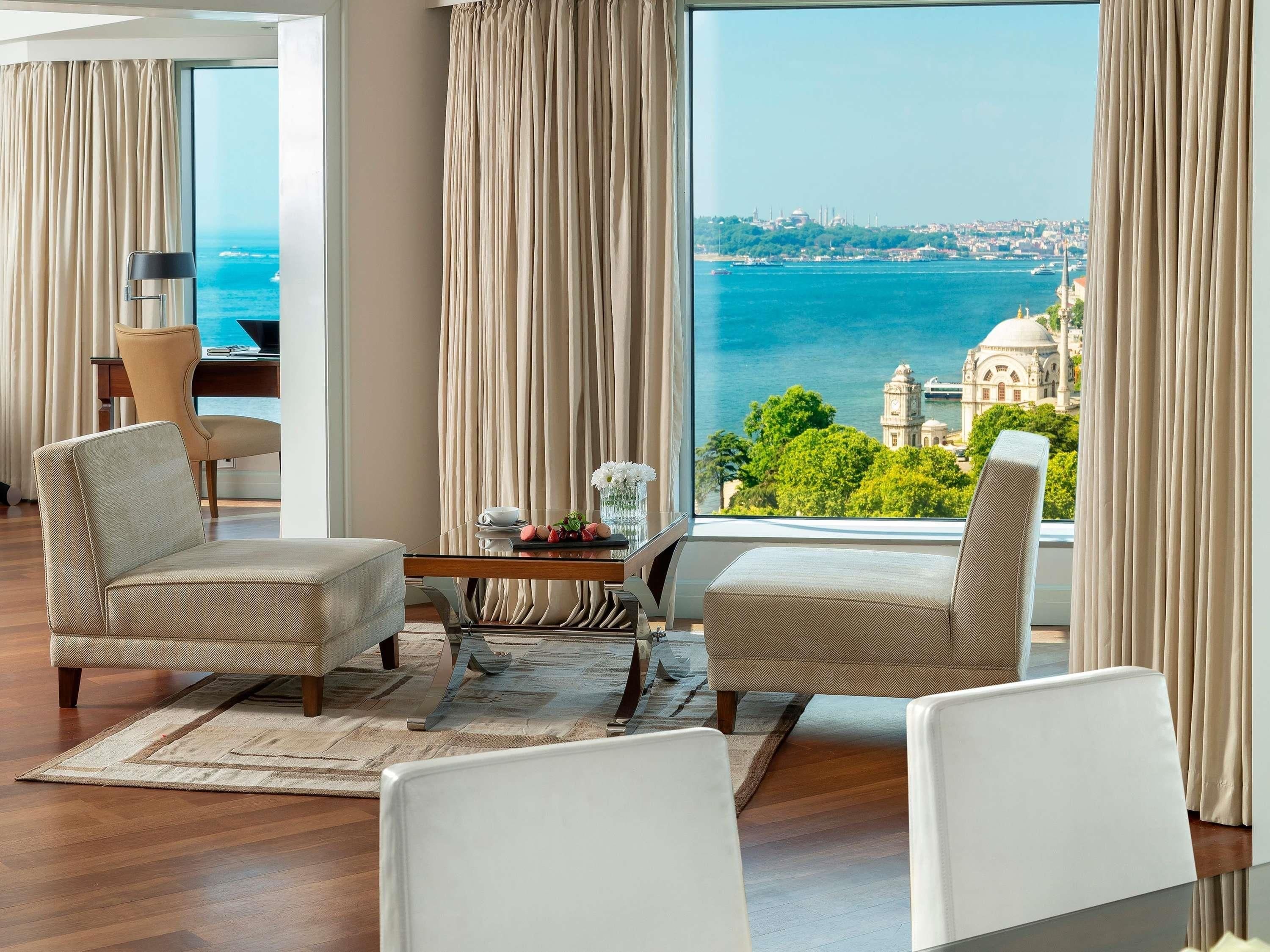 A modern living room with a large window, two armchairs, a small table with a vase and a cup, and a view of a cityscape and sea.