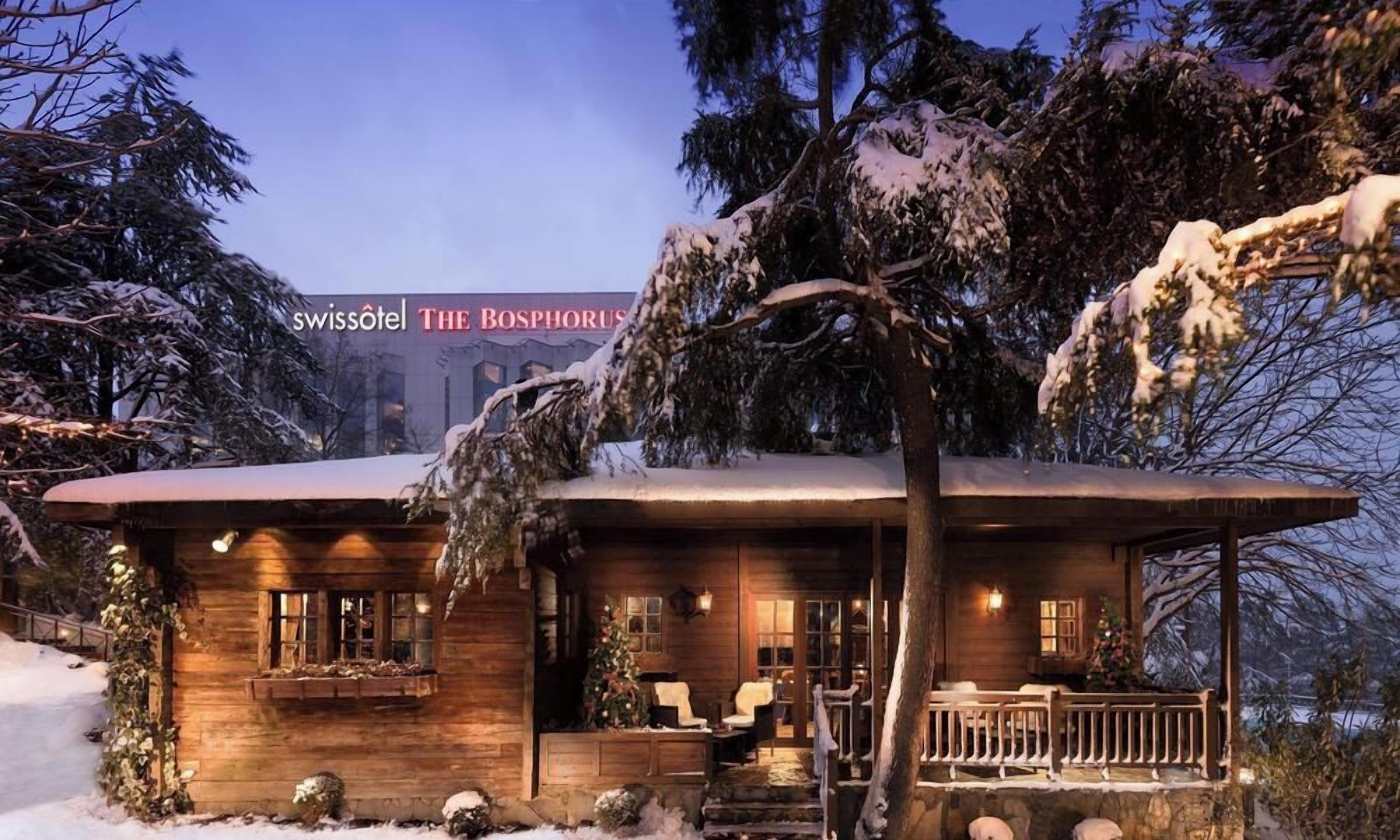Una cabaña de madera con un porche, un árbol de Navidad y una vista del hotel The Bosphorus al fondo.
