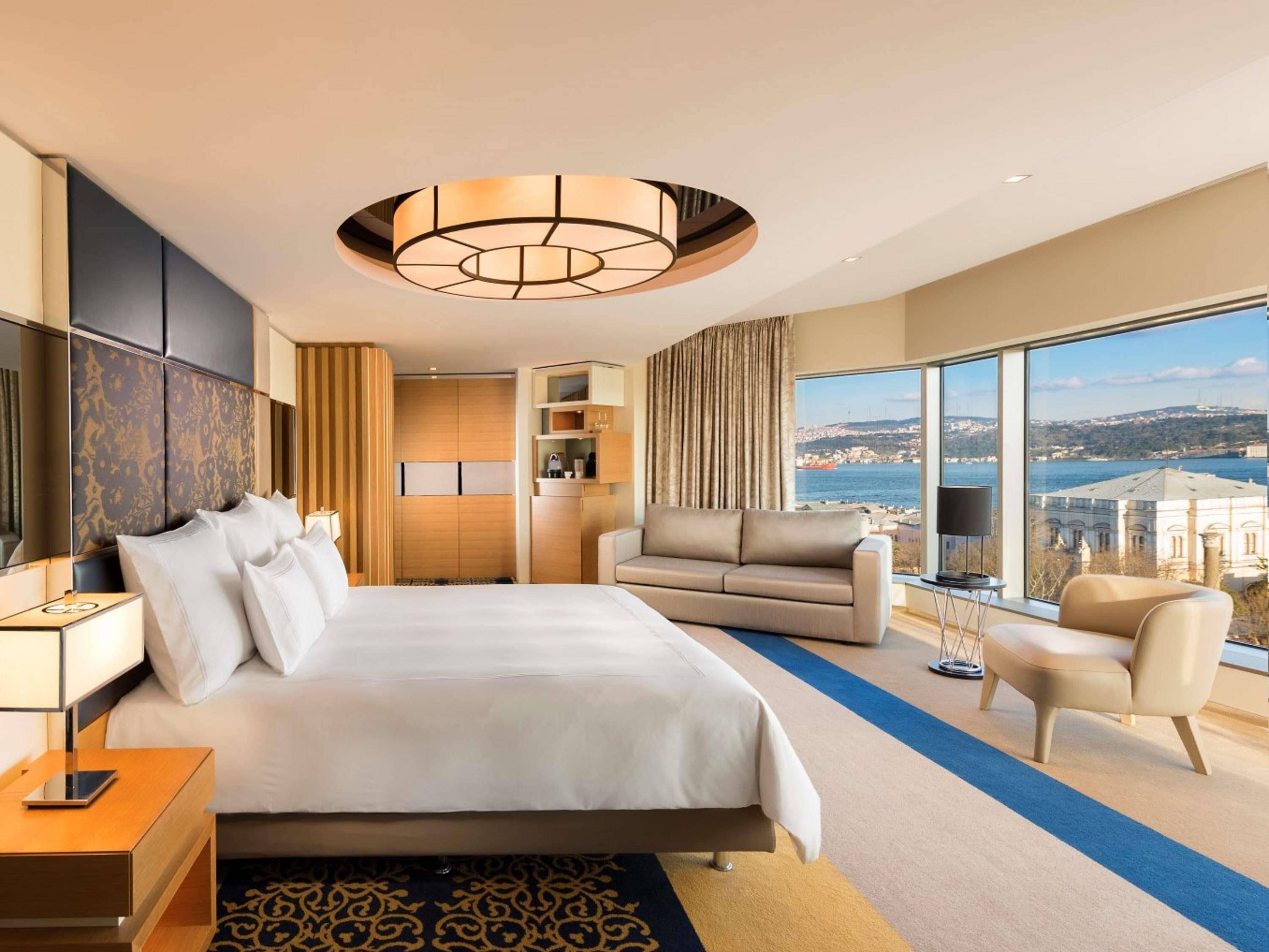 A hotel room with a large bed, a sofa, a desk, and a view of a cityscape.