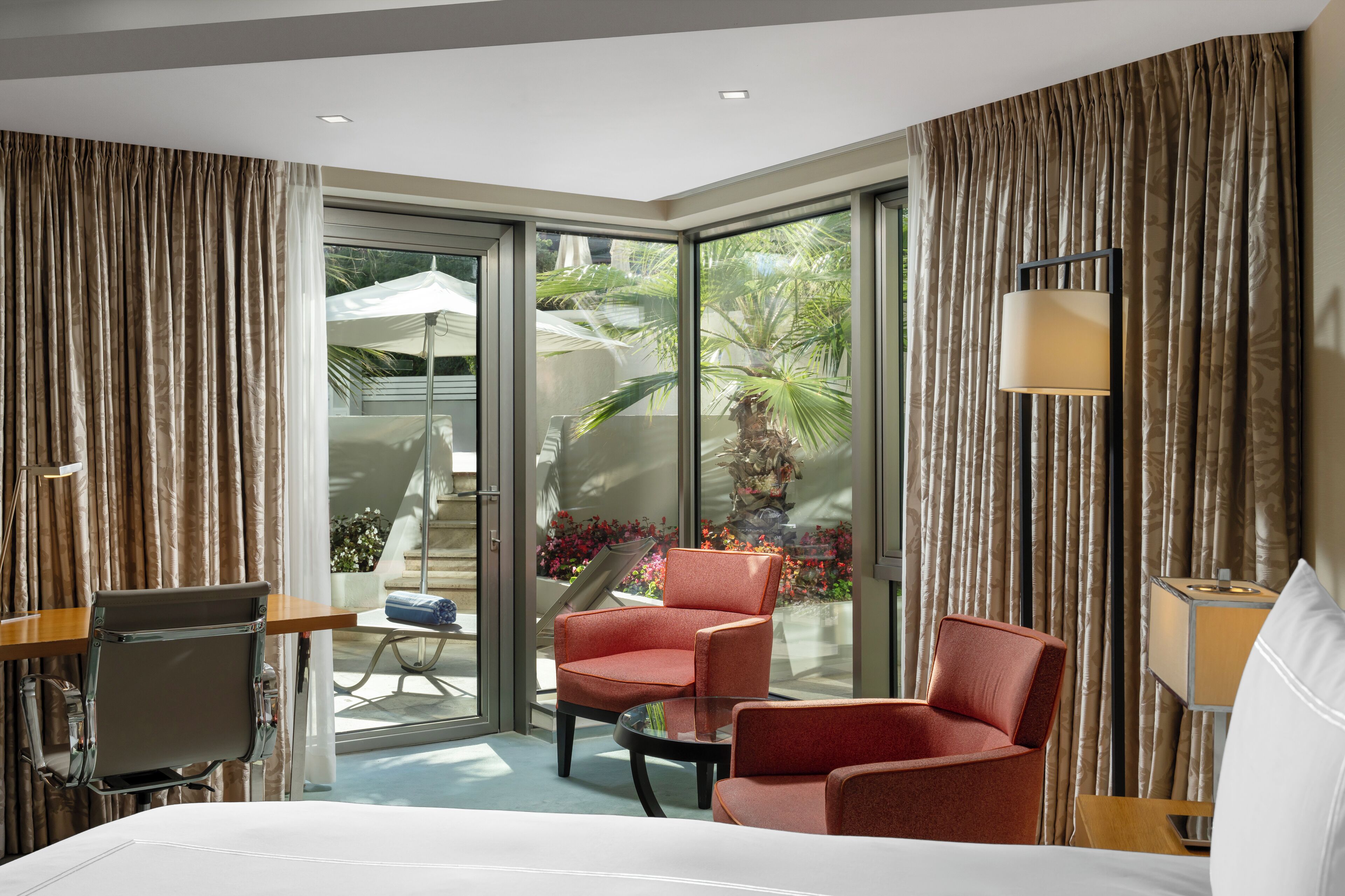 Habitación de hotel con cama, dos sillas rojas, una mesita, escritorio, lámpara y vista a un jardín a través de puertas de vidrio grandes.
