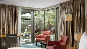 Habitación de hotel con cama, dos sillas rojas, una mesita, escritorio, lámpara y vista a un jardín a través de puertas de vidrio grandes.