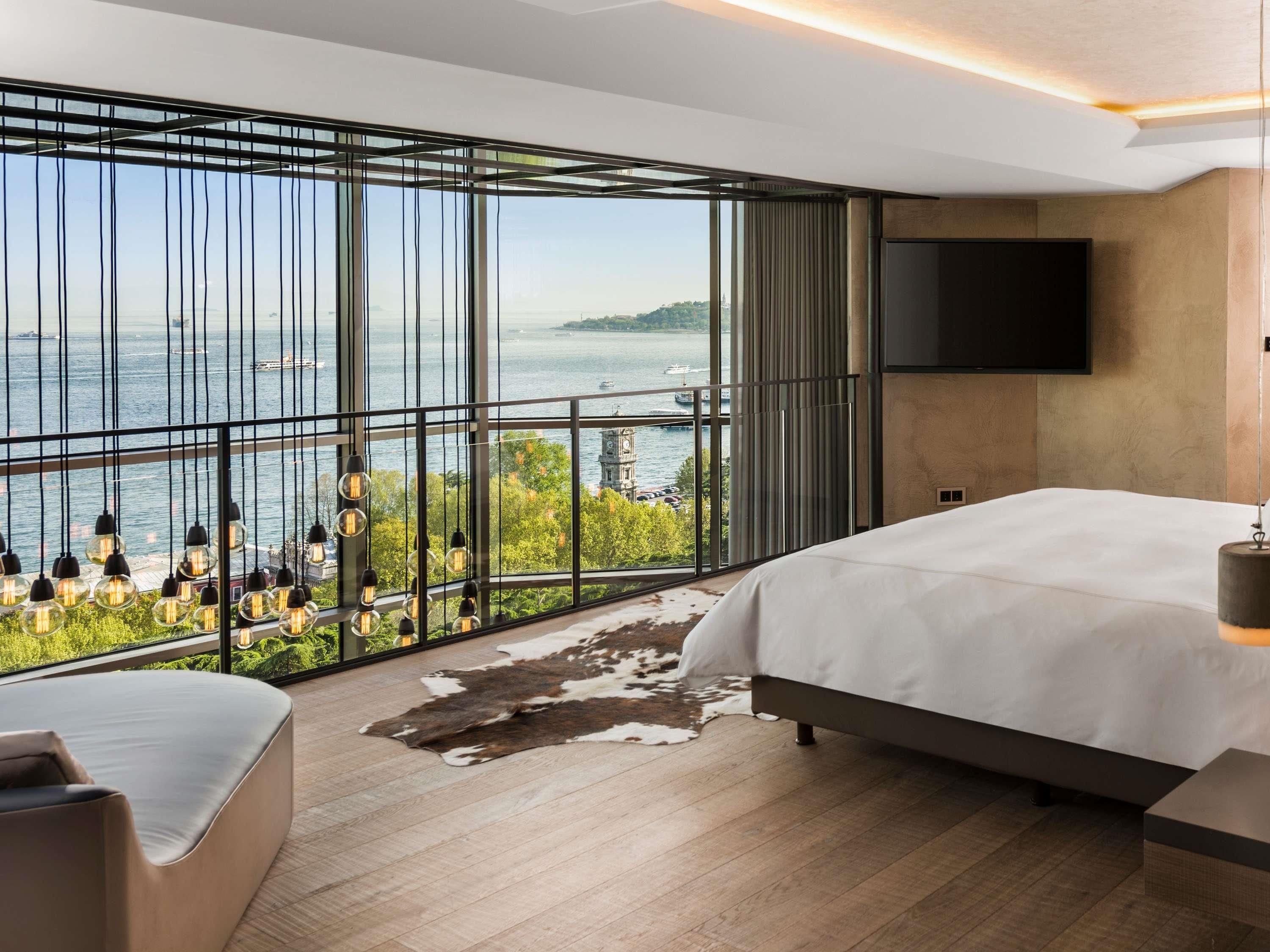 A modern bedroom with a large window, a bed, a TV, and a view of the water.