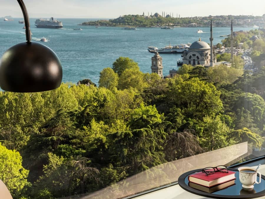 Une chambre située dans un immeuble de grande hauteur, offrant une vue sur une étendue d’eau, un paysage urbain lointain, ainsi qu’un coin repas avec une table, des livres et une tasse.