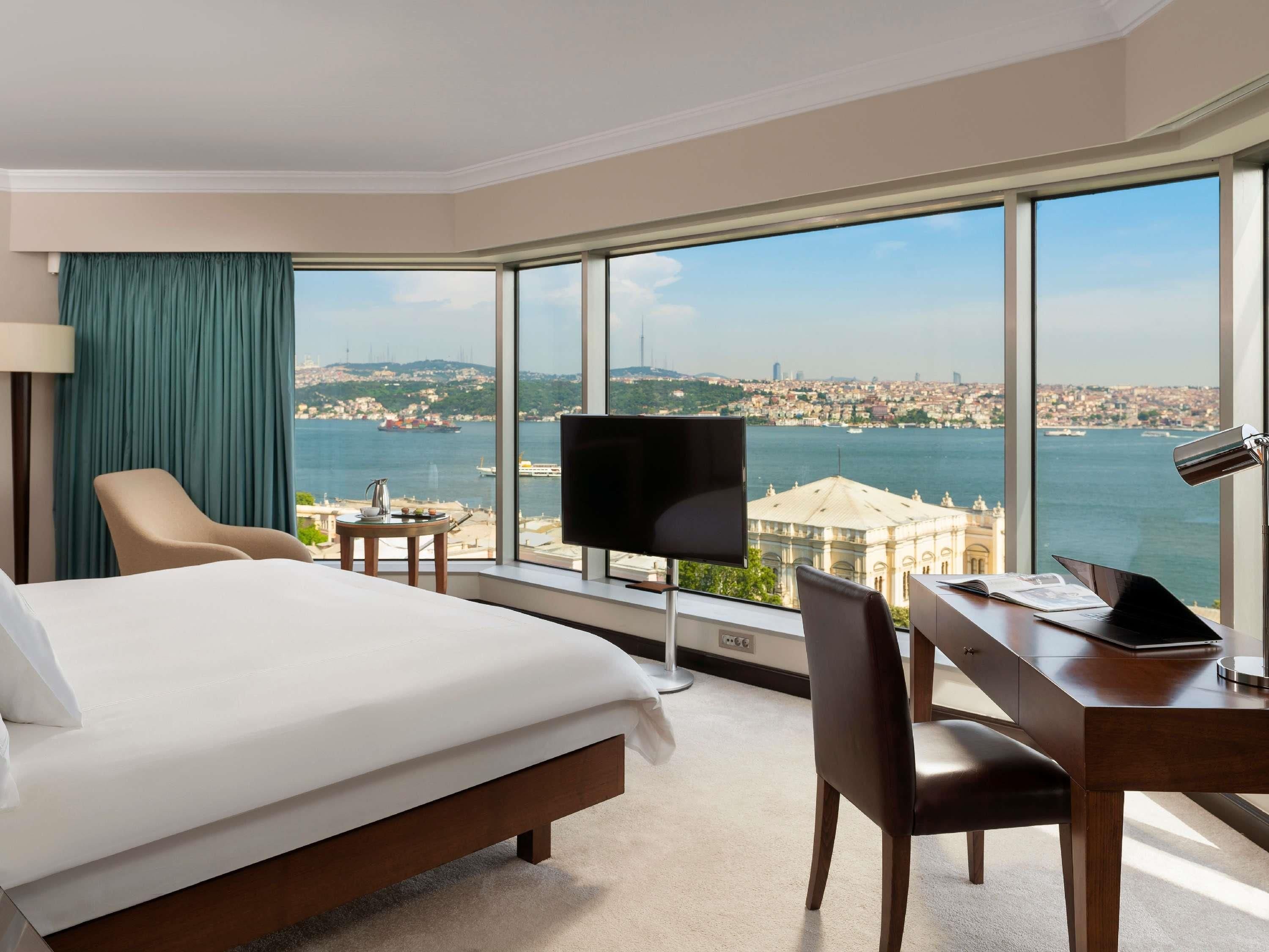 A hotel room with a large bed, a desk, a chair, a TV, and a view of a city and body of water.
