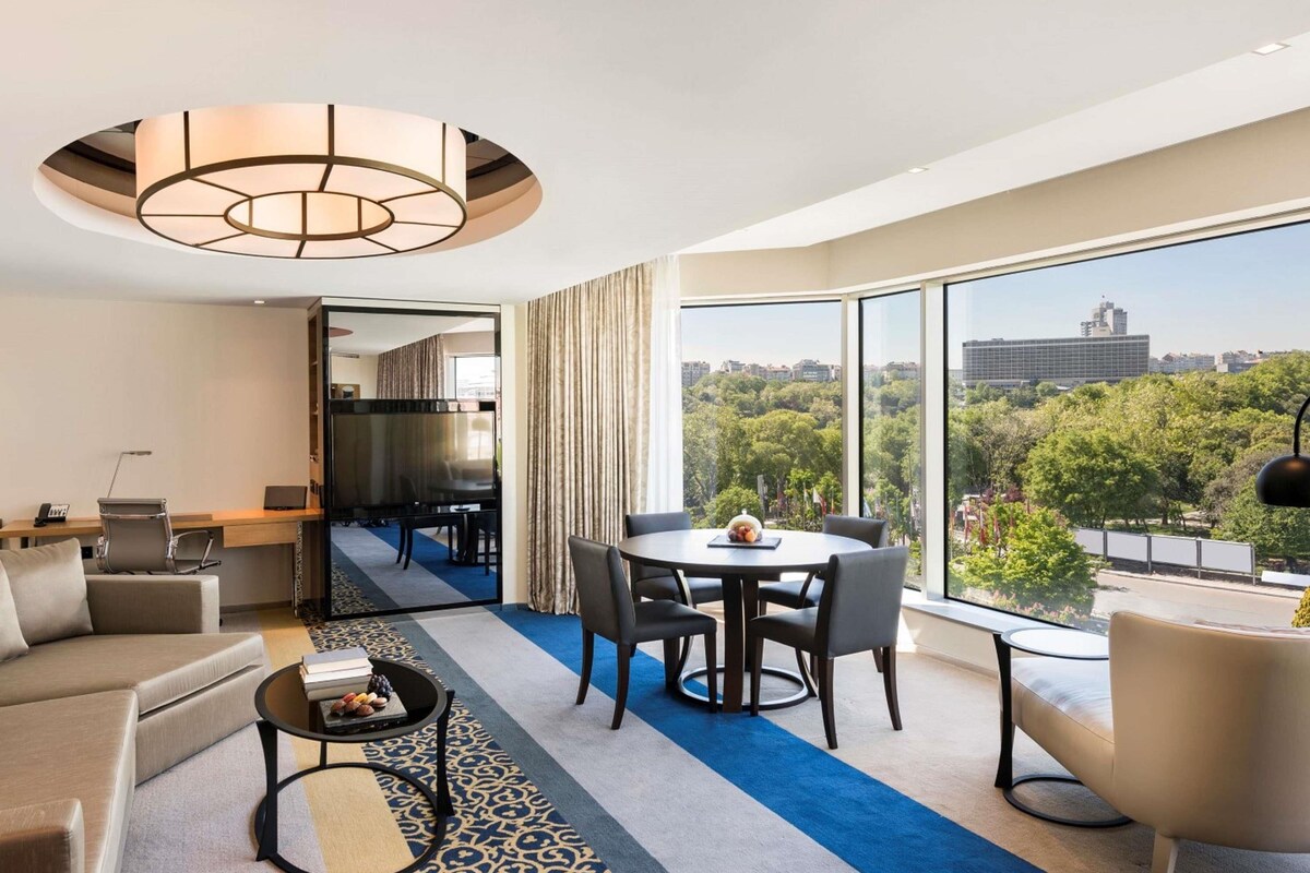 A modern hotel room with a large window, a dining area, a sofa, a coffee table, and a desk.