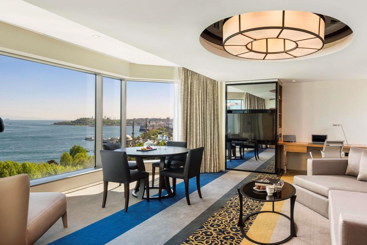 A modern hotel room with a large window offering a city view, a dining area with a table and chairs, a sofa, a coffee table, and a television.