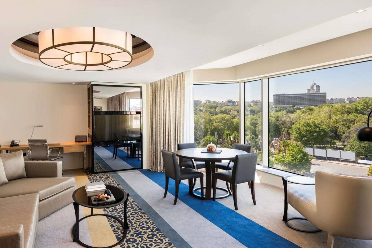 A modern hotel room with a large window, a dining area, a sofa, a coffee table, and a desk.
