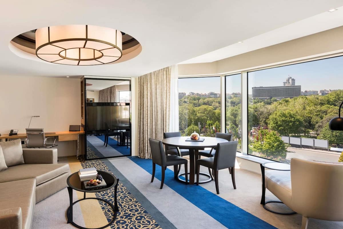 Una habitación de hotel moderna con un ventanal grande, una zona de comedor, un sofá, una mesa de centro y un escritorio.