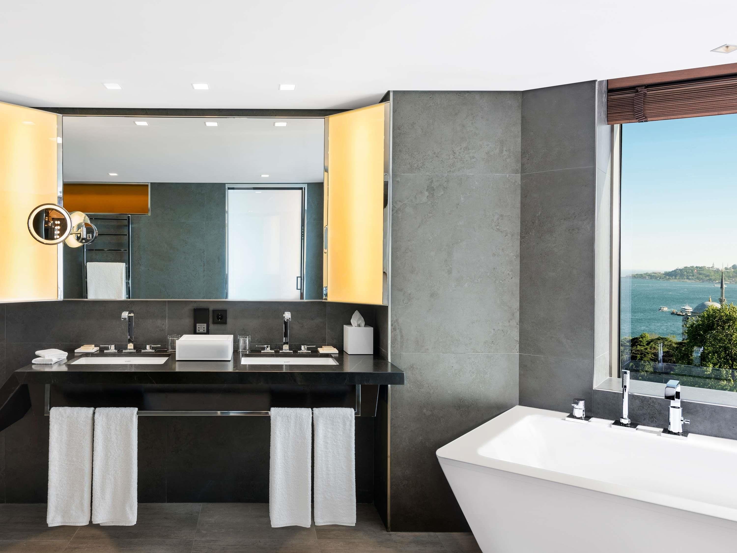 A modern bathroom with a large mirror, two sinks, a bathtub, and a view of a body of water.