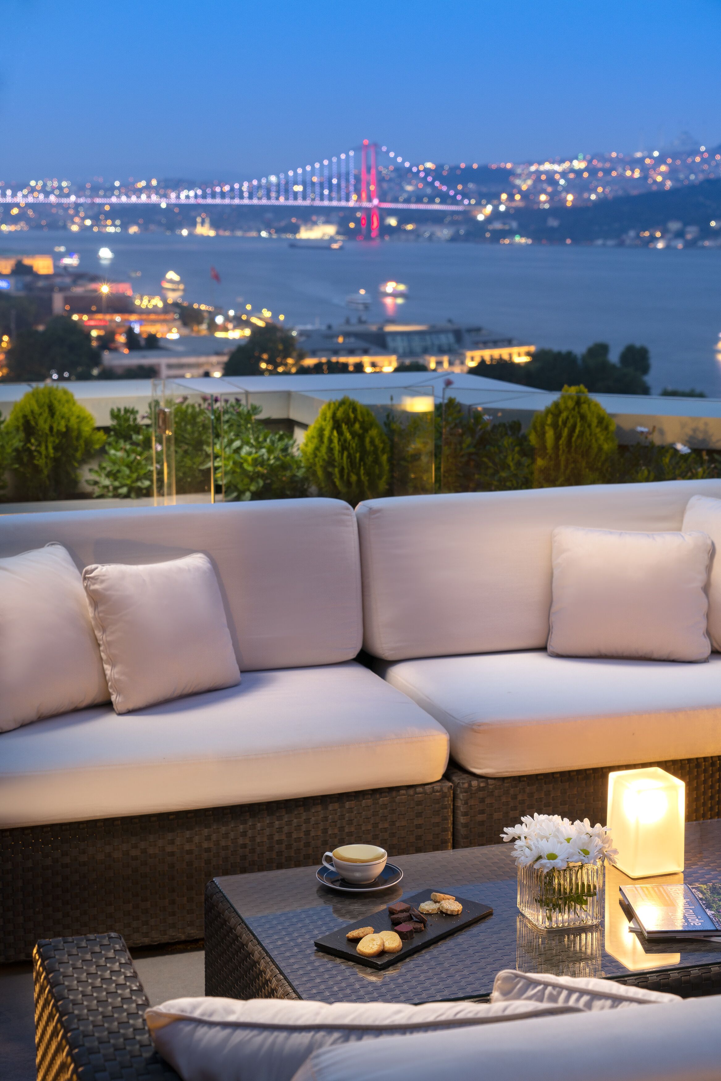 A modern outdoor seating area with a sofa and a table, overlooking a cityscape with a bridge and waterfront.