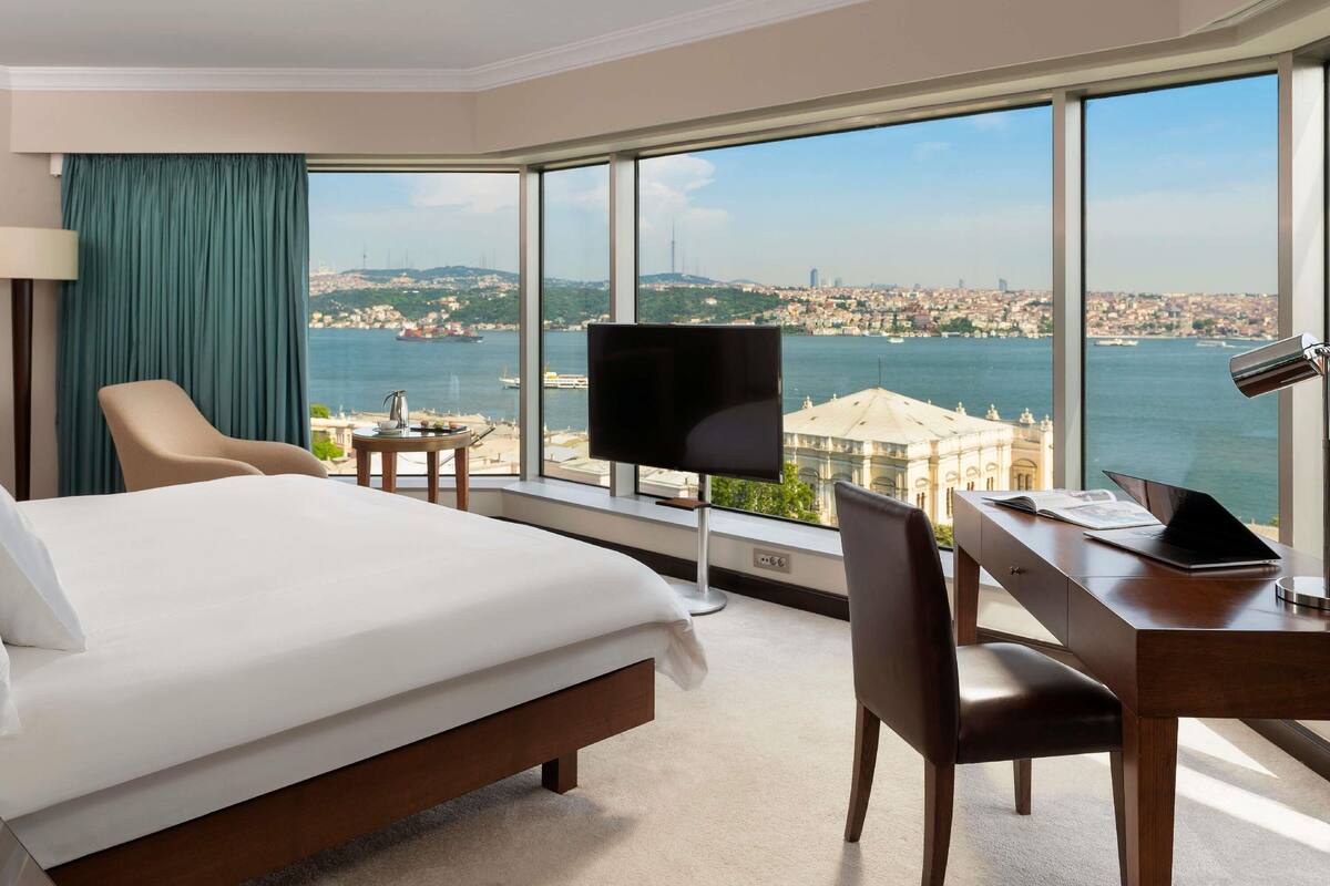 A hotel room with a bed, desk, chair, TV, and large windows with a city and water view.