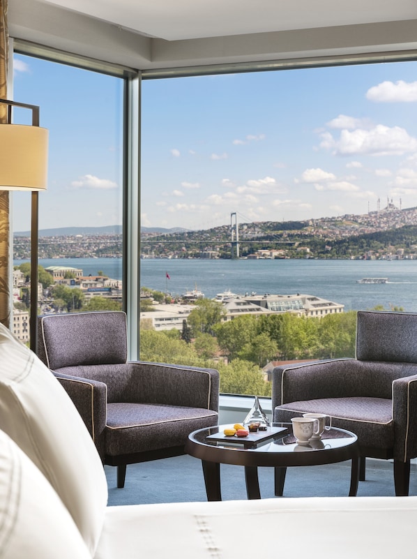 A modern room with a large window offering a view of a bridge and cityscape. There are two upholstered chairs and a small round table with a teapot and pastries. - Swissotel The Bosphorus Istanbul (Istanbul)
