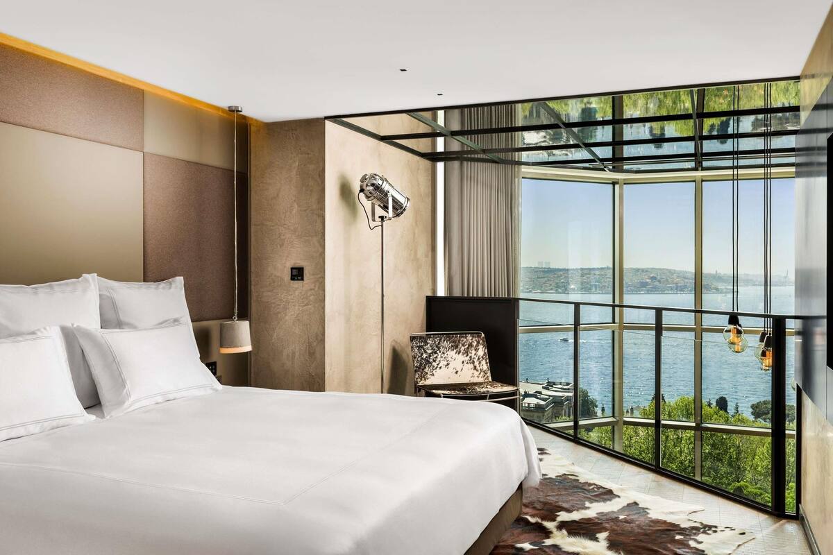 A modern hotel room with a large bed, a view of a body of water, a leather rug, and a unique wall-mounted light fixture.
