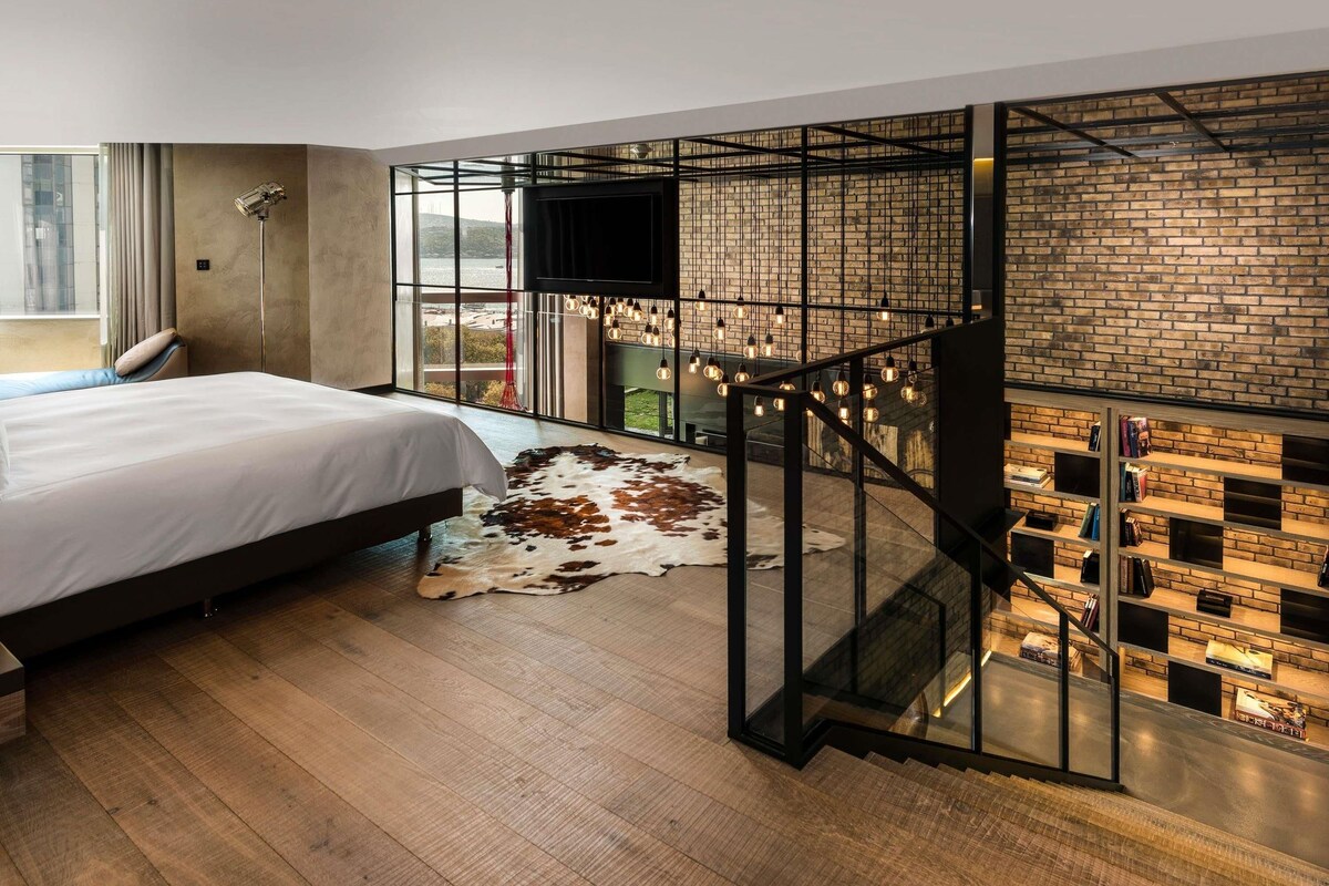 A modern hotel room with a large bed, a leather sofa, a glass staircase, a brick wall, and a TV mounted on the wall.
