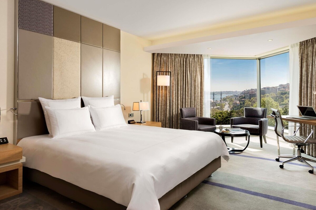 A hotel room with a large bed, a desk with a laptop, a chair, and a view of a cityscape.