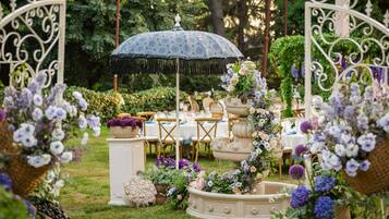 An outdoor garden setting with a decorative fountain, tables, and chairs arranged for an event.