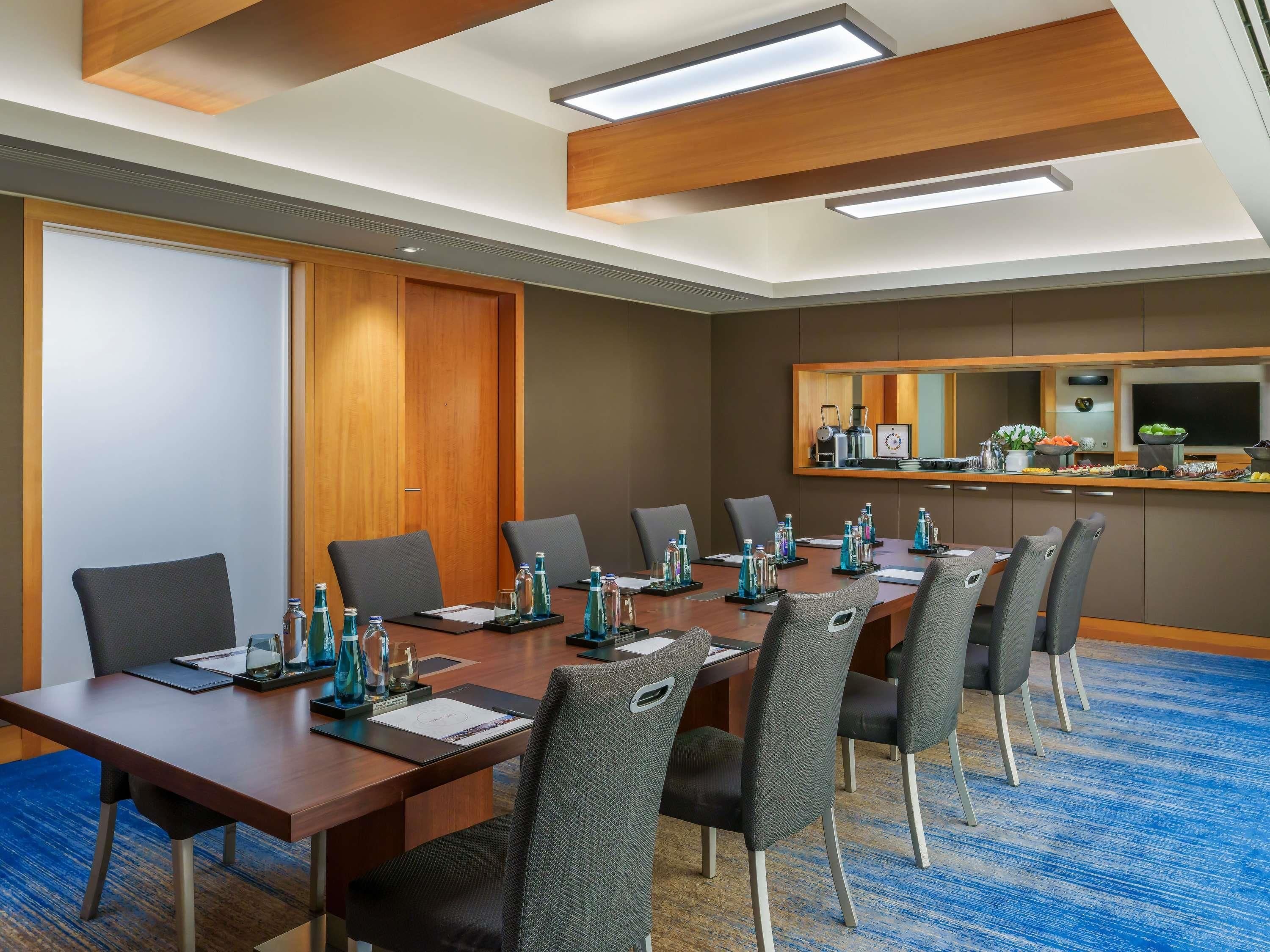 A conference room with a long table, chairs, a wooden door, a mirror, and a kitchenette area.