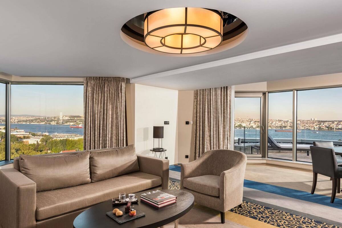 A modern hotel room with a sofa, armchair, and a table with snacks. It features large windows with a city and water view.