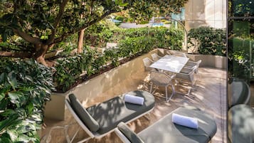 Un área de estar al aire libre iluminada por el sol, con una mesa y sillas, rodeada de abundante vegetación y un gran árbol.