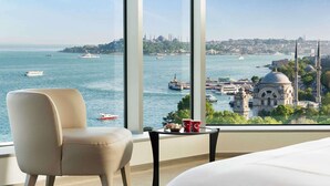 A hotel room with a large window offering a view of a cityscape, including a prominent mosque and a body of water with boats.