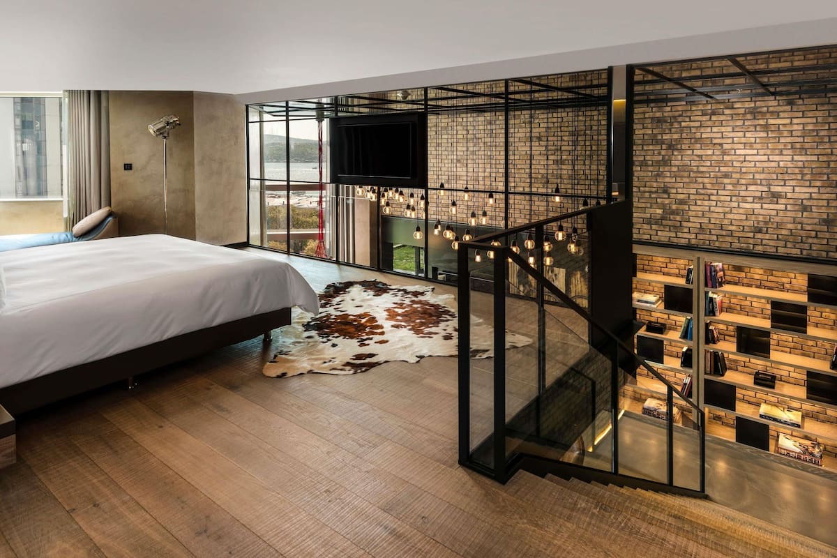 A modern hotel room with a large bed, a leather sofa, a glass staircase, a brick wall, and a TV mounted on the wall.
