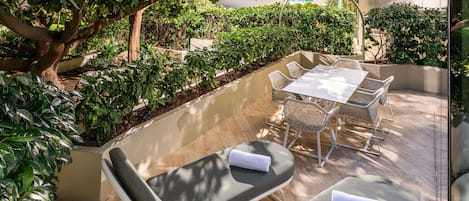 An outdoor patio with a table and chairs, surrounded by greenery and a tree.