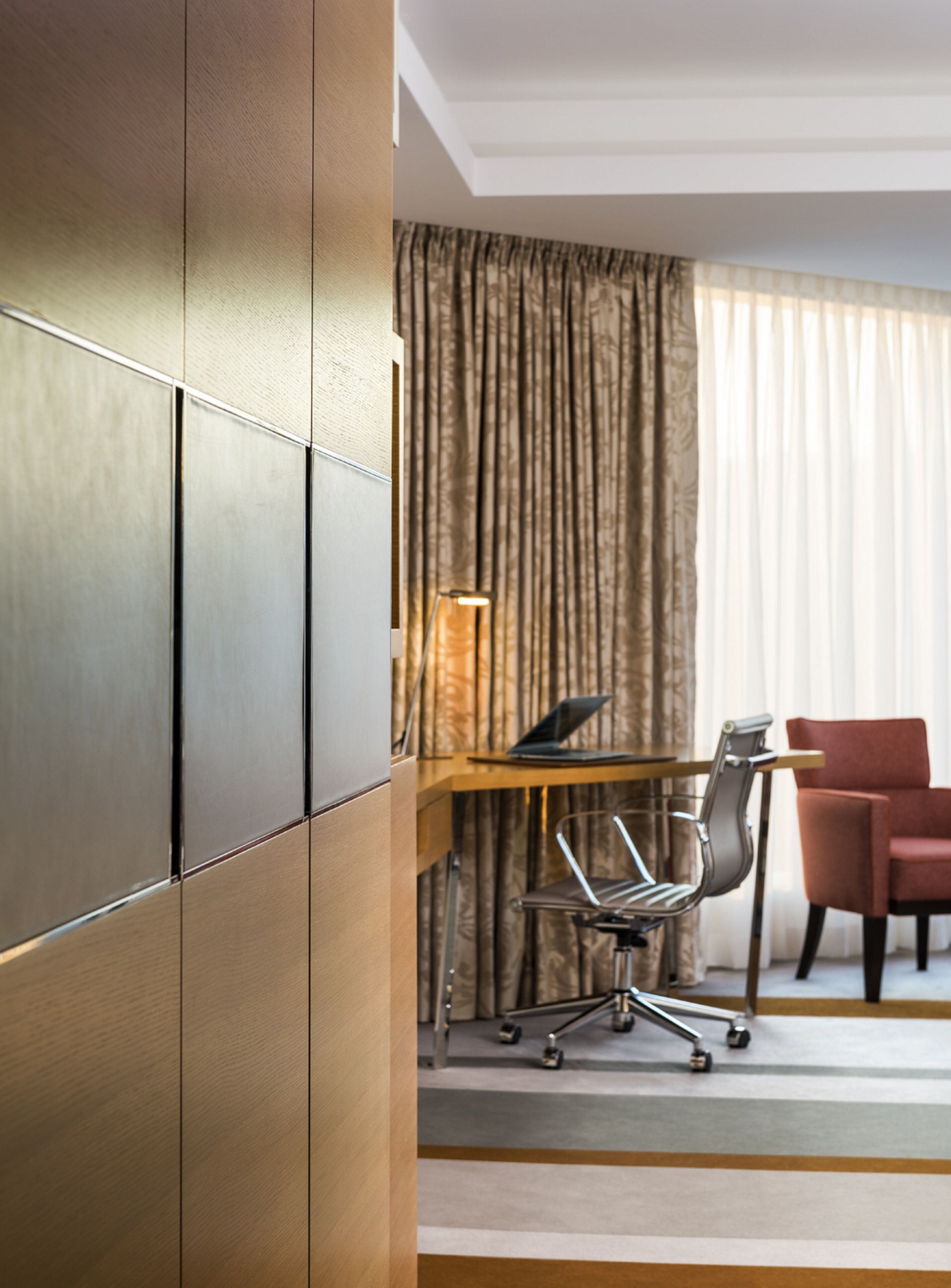 Ein Hotelzimmer mit Schreibtisch aus Holz, einem Laptop, einem modernen Stuhl und einem roten Sessel.