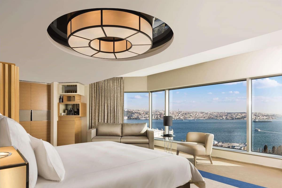 A modern hotel room with a large bed, a seating area with two chairs, a desk, and a view of a cityscape and water.
