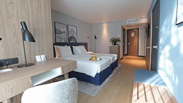 Una habitación de hotel moderna con un escritorio de madera, una cama con detalles azules, una silla blanca y una mesita de noche con un jarrón de flores.