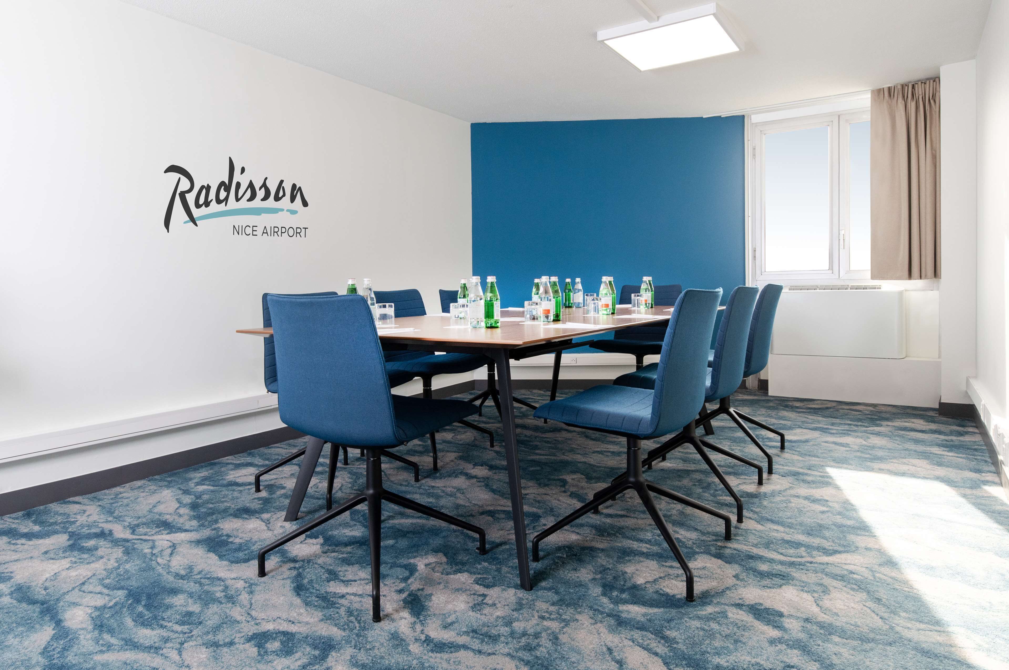 Ein Konferenzraum mit blauem Teppich, einer weißen Wand mit dem Radisson-Logo und einem großen Fenster mit beigen Vorhängen.