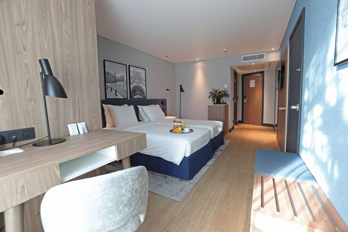 A modern hotel room with a wooden desk, two beds, a bedside table, a chair, and a small desk lamp.