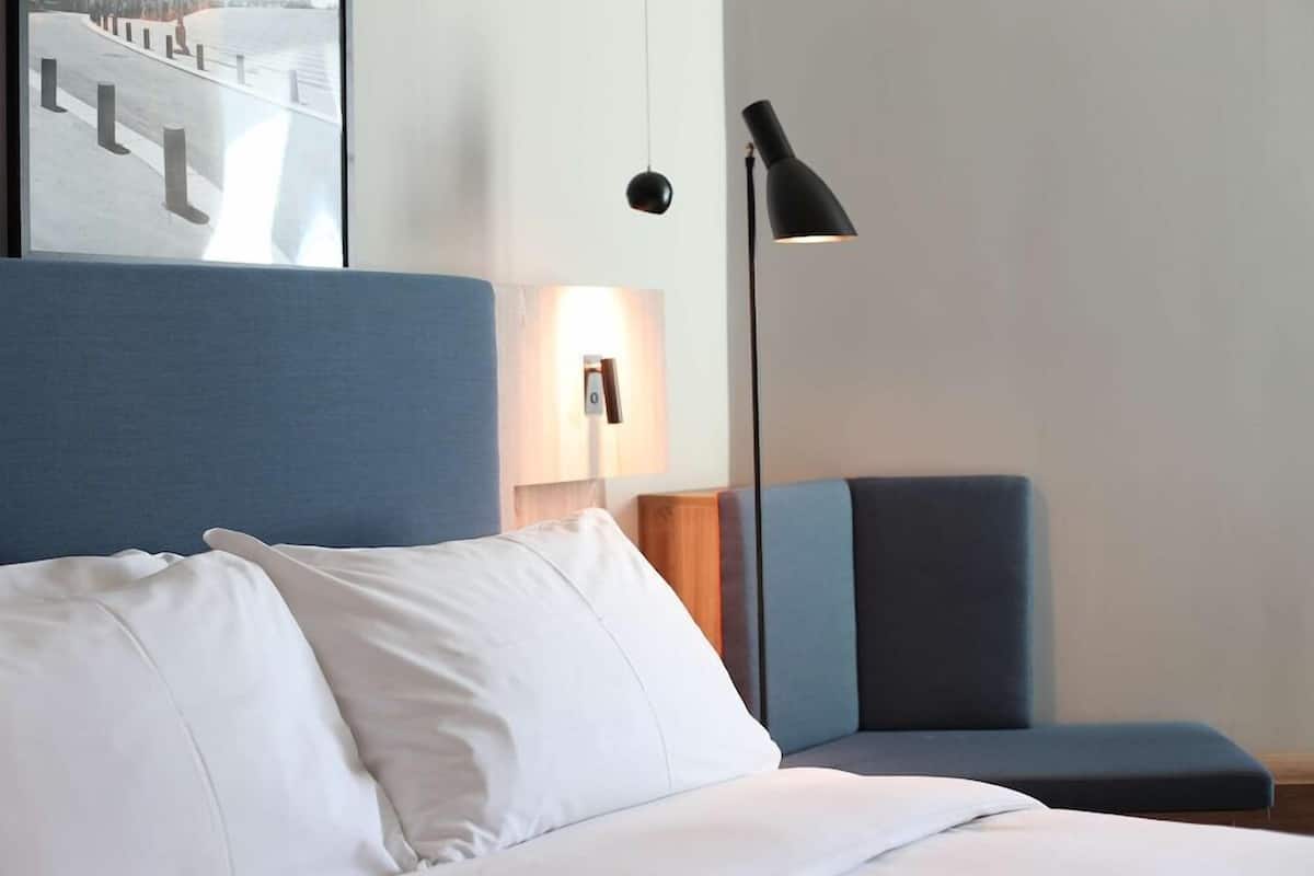 A hotel room with a bed, a blue armchair, a standing lamp, and a wall-mounted light fixture.