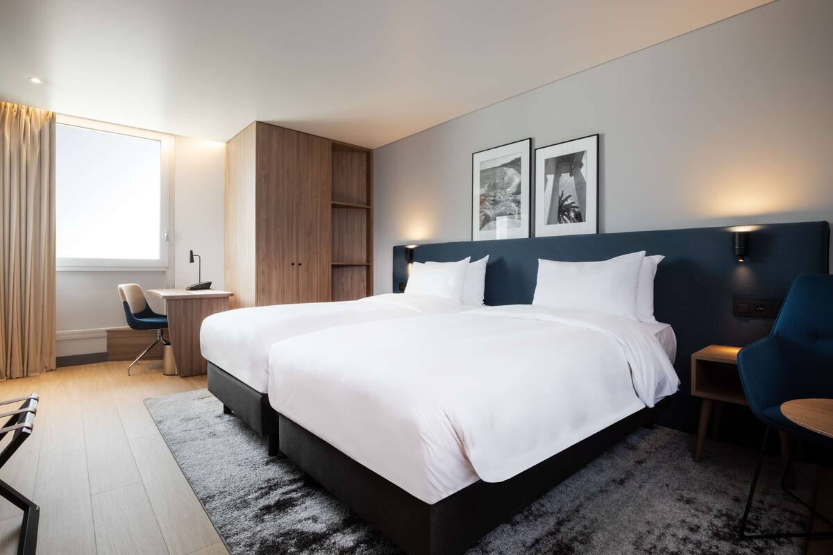 A modern hotel room with a large bed, a desk, and a chair. There are two framed pictures on the wall, a window with curtains, and a carpeted floor.