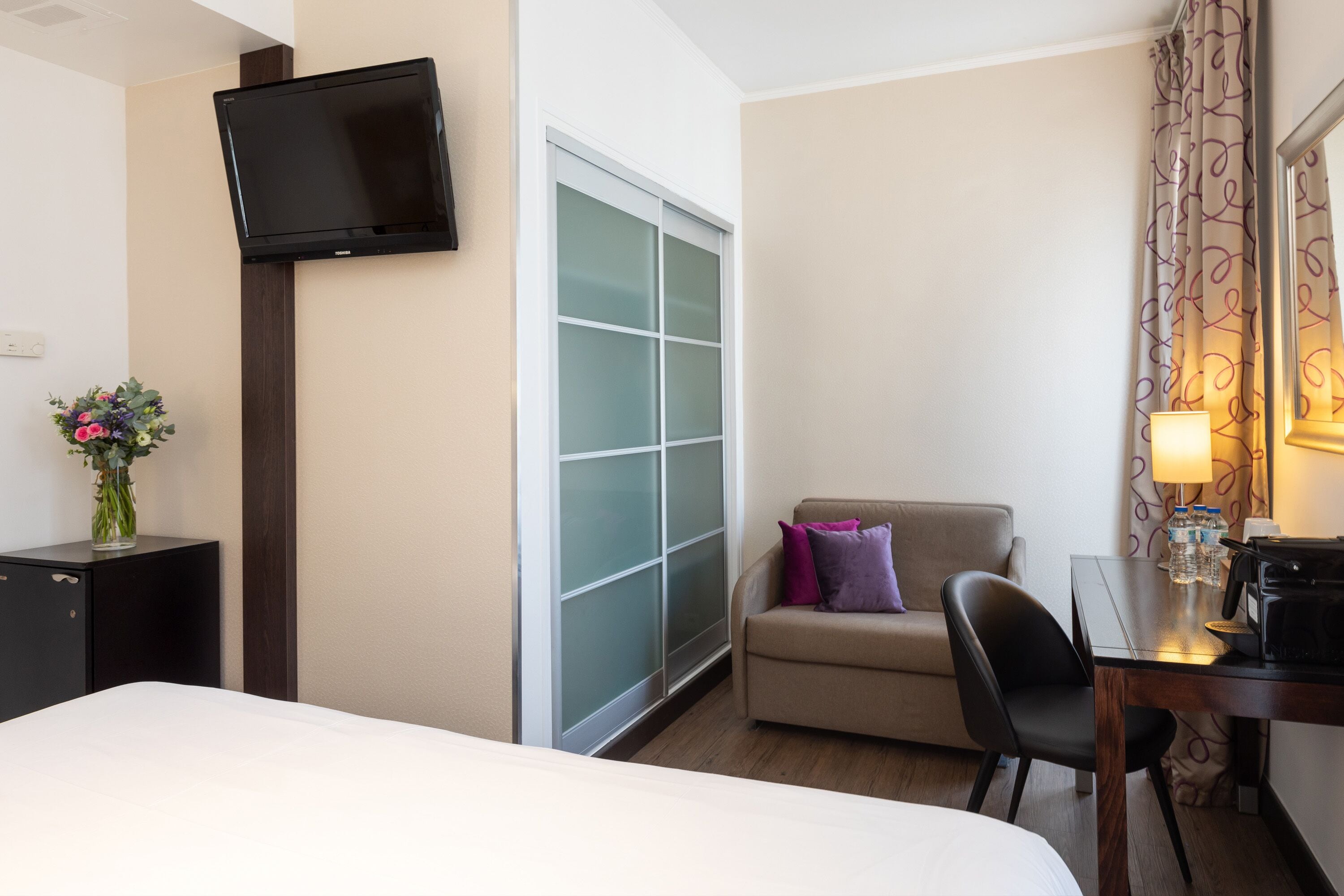 A hotel room with a bed, a desk with a lamp, a TV mounted on the wall, a small sofa, and a window with frosted glass.