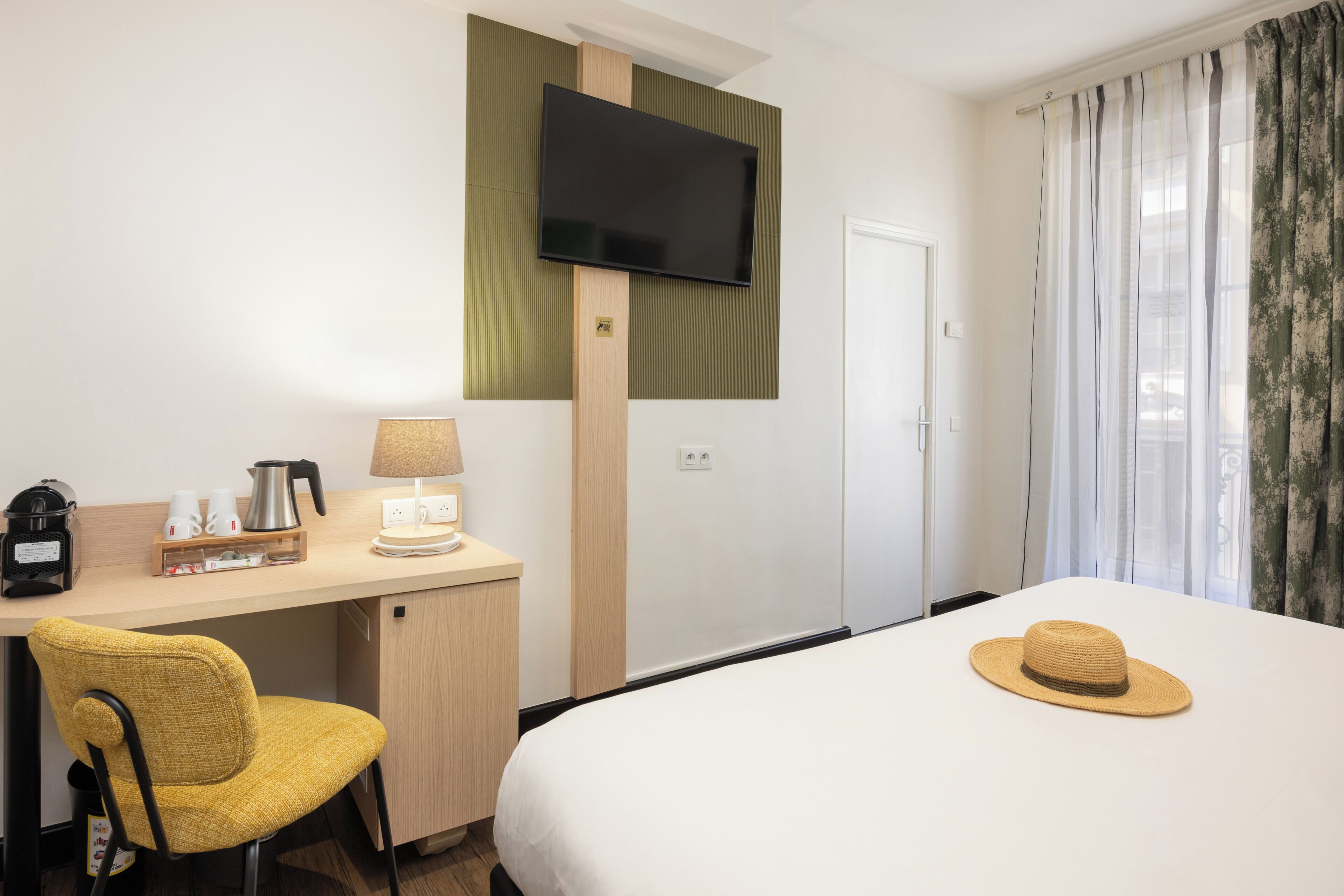 A hotel room with a bed, a desk with a lamp, a chair, a television mounted on the wall, and a window with curtains.