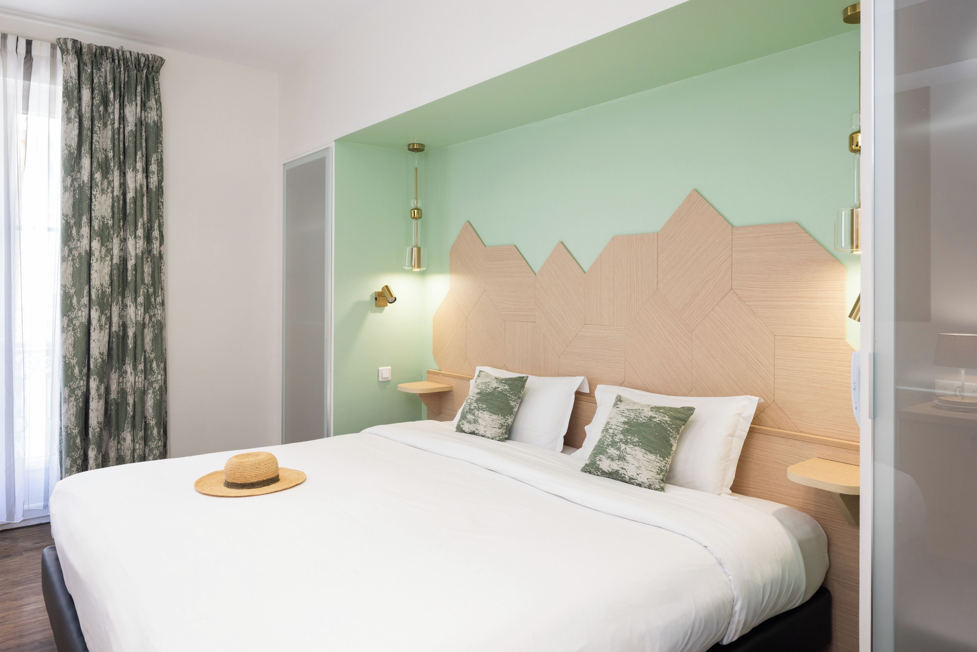 Hotellihuone, jossa on sänky, sängyllä hattu, yöpöytä, seinään kiinnitetty valaisin ja verho.