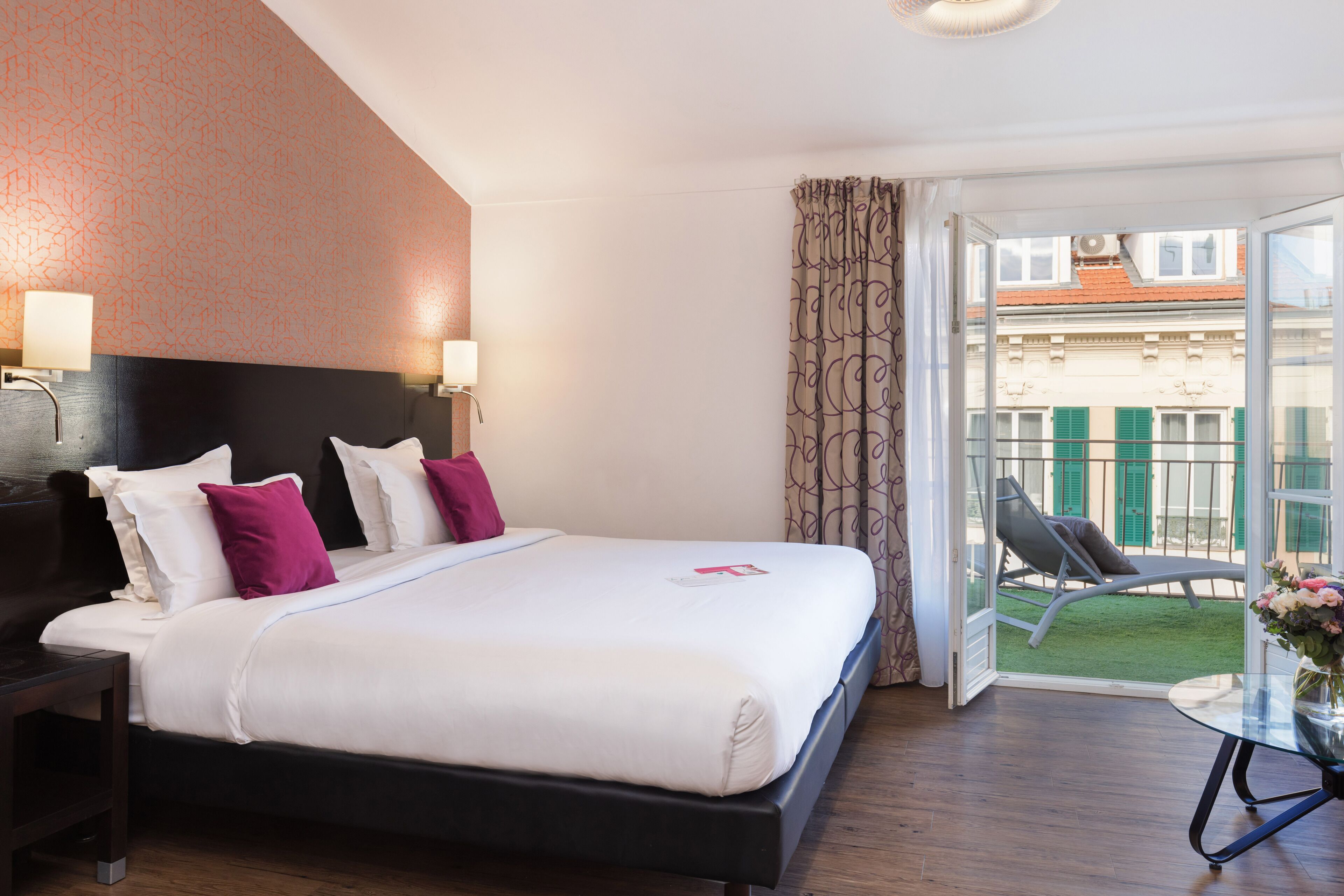 Une chambre d’hôtel avec un lit, des lampes de chevet, une petite table avec un vase et un balcon avec une chaise.