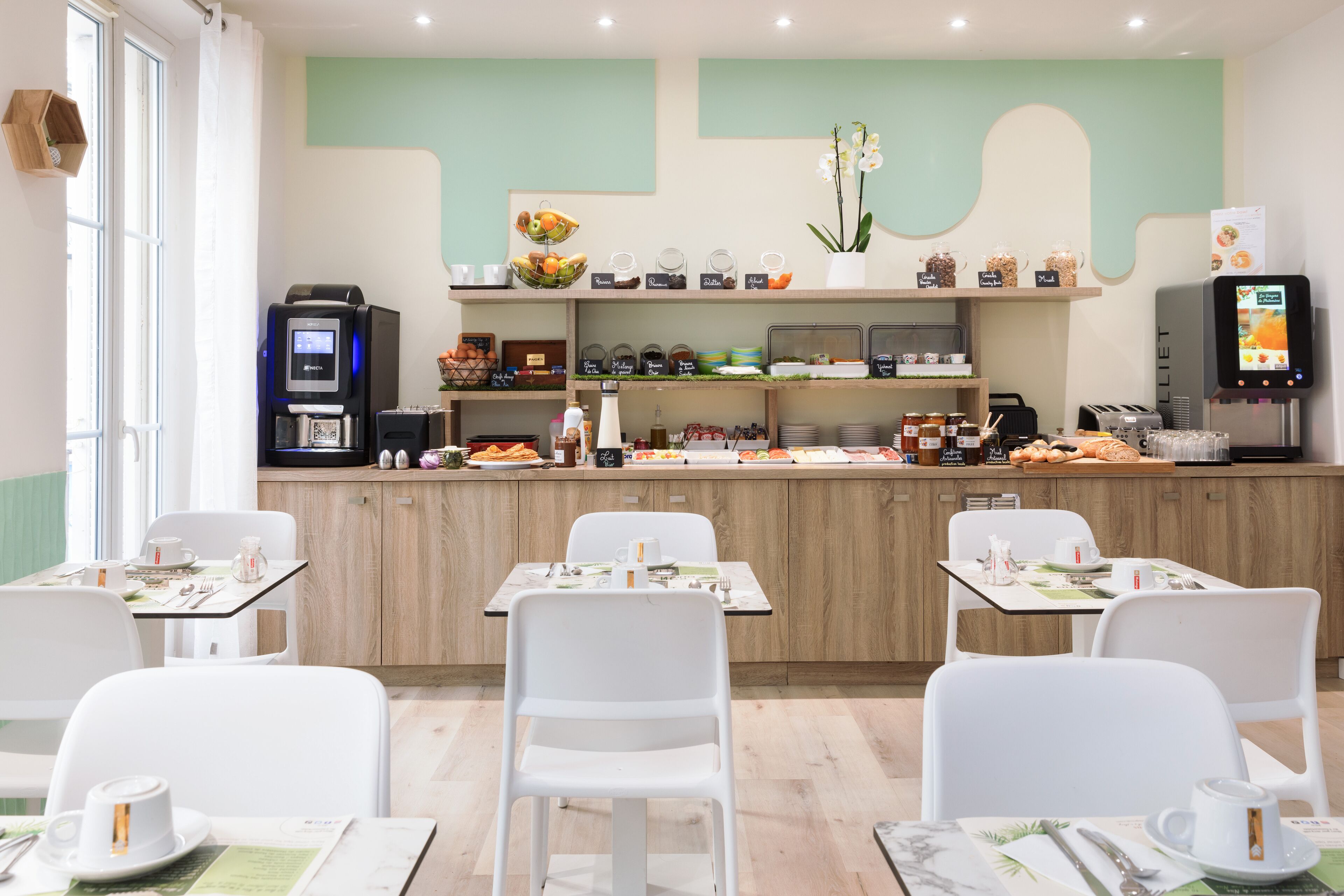 Zona de desayuno con mostrador que exhibe diferentes alimentos y una máquina de café. Hay sillas y mesas blancas dispuestas para comer.