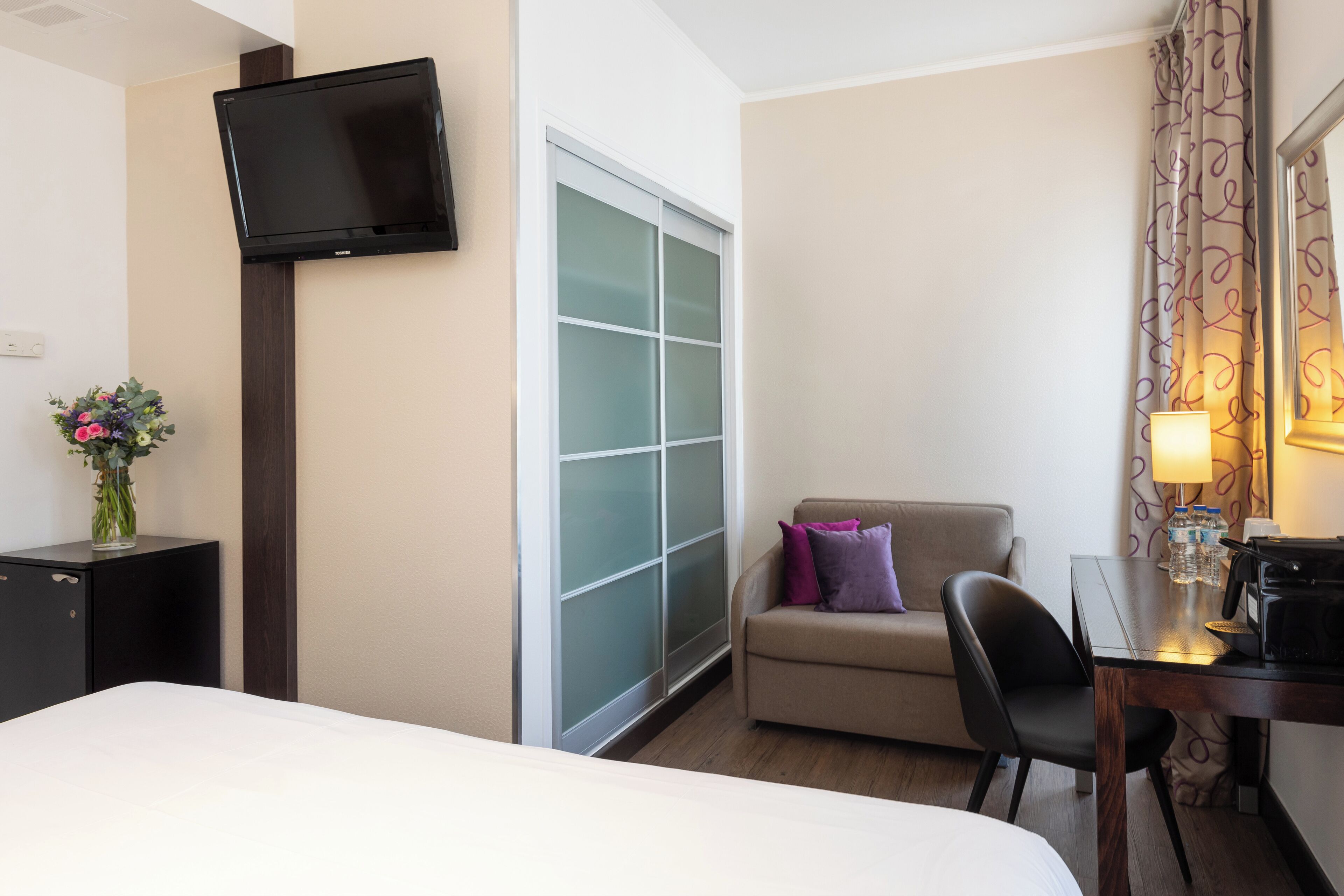 A hotel room with a bed, a desk with a lamp, a TV mounted on the wall, and a window with frosted glass panels.
