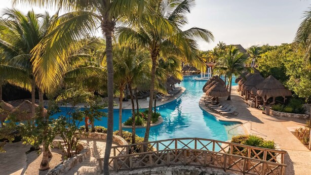A resort pool area with palm trees, lounge chairs, and a stone bridge.