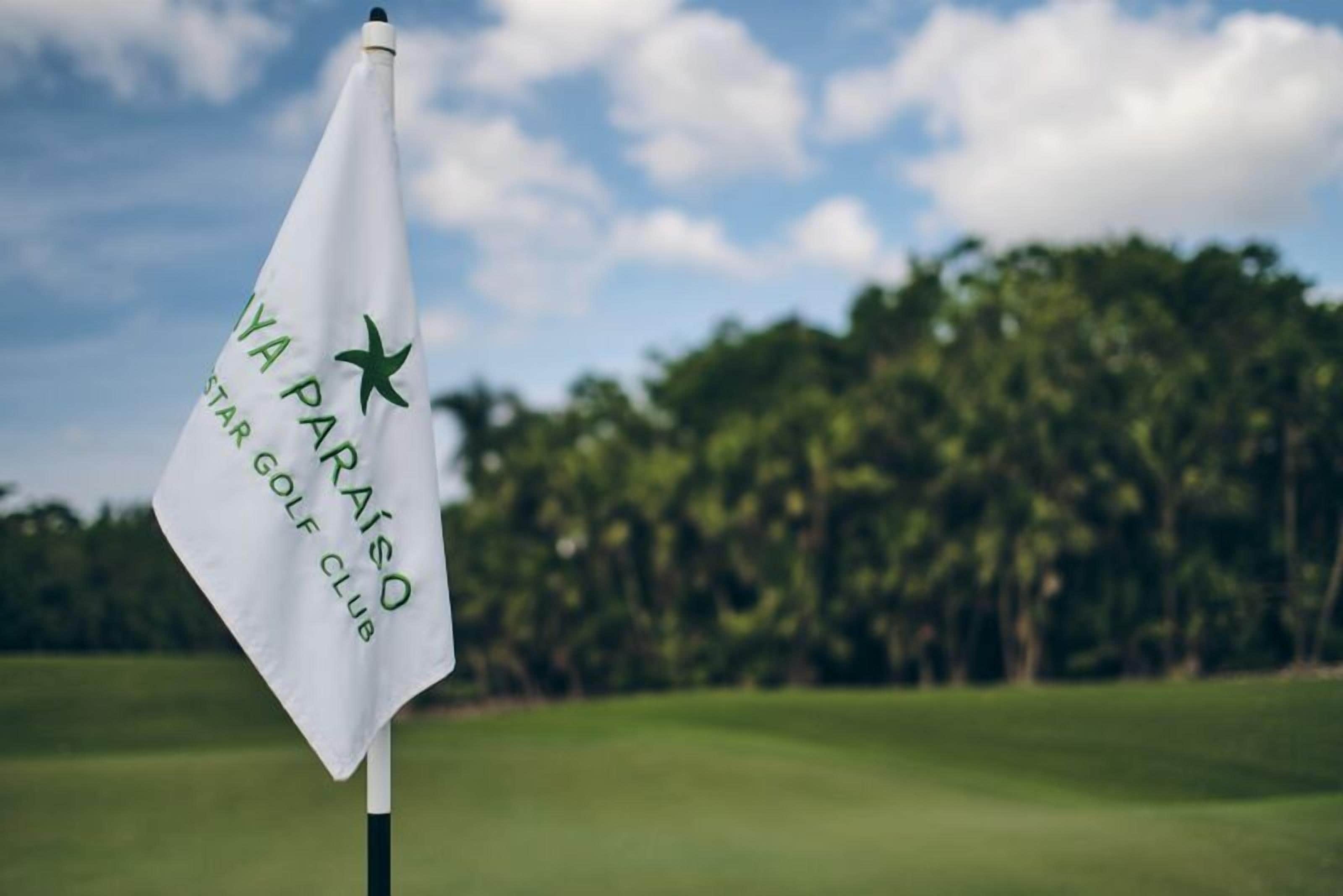 Una bandera de golf con la inscripción "IYA PARAISO" y "STAR GOLF CLUB", sobre un fondo de campo de golf y árboles.
