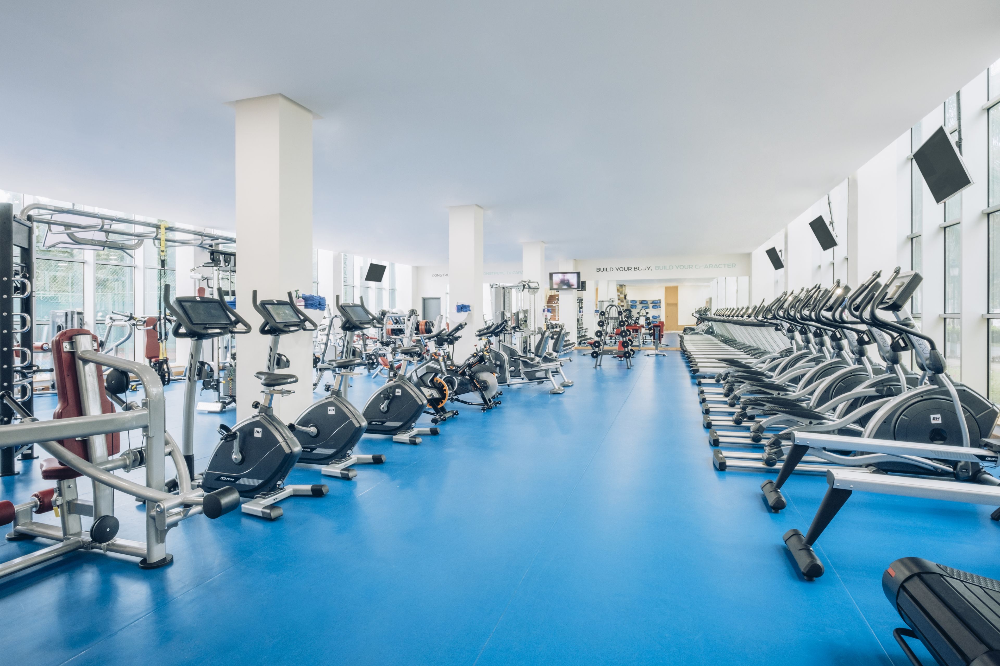 Ein Fitnessstudio mit verschiedenen Trainingsgeräten, einem blauen Boden und großen Fenstern.