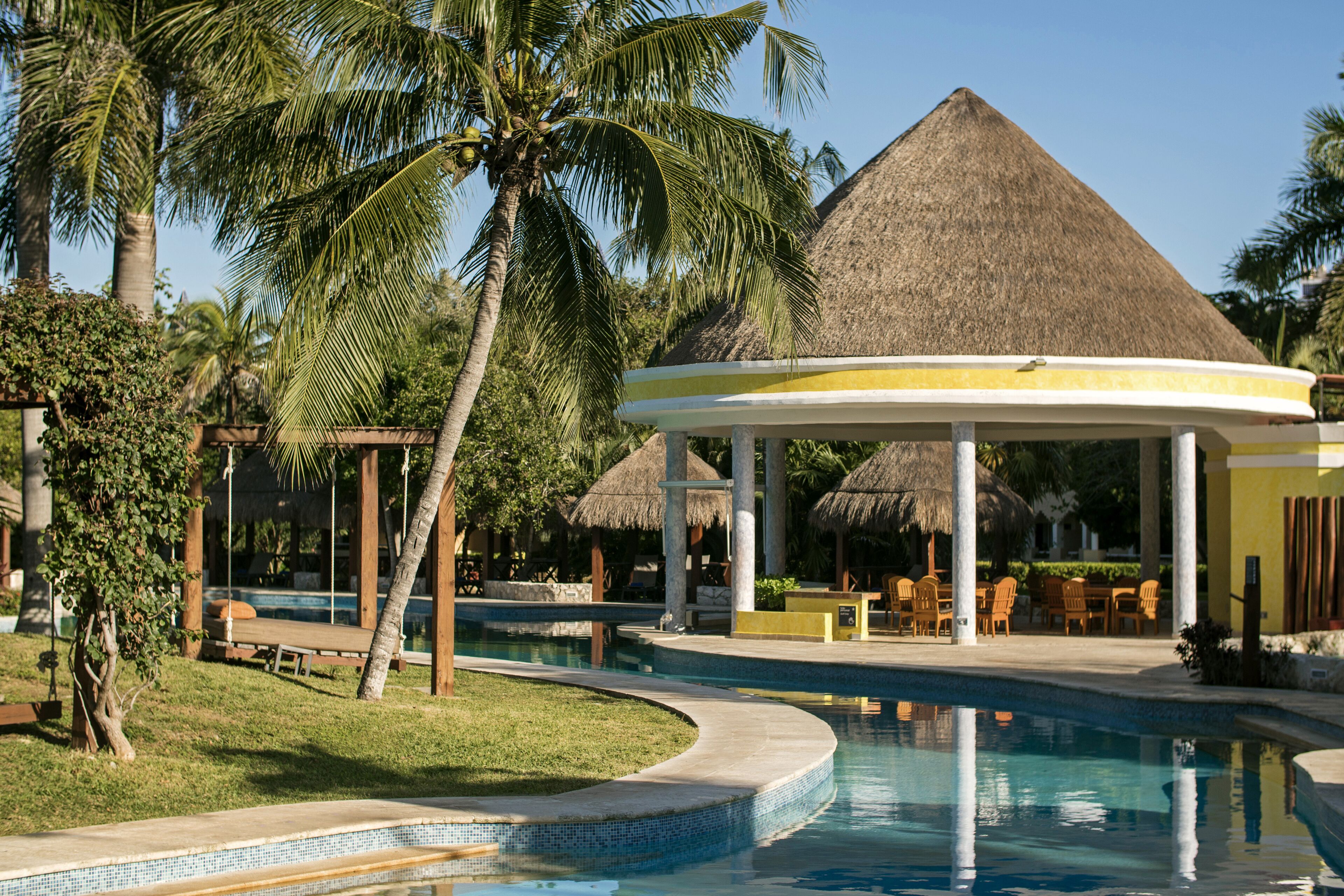 Un espace piscine aménagé dans un complexe hôtelier, avec un gazebo à toit de chaume, des palmiers et des chaises longues.