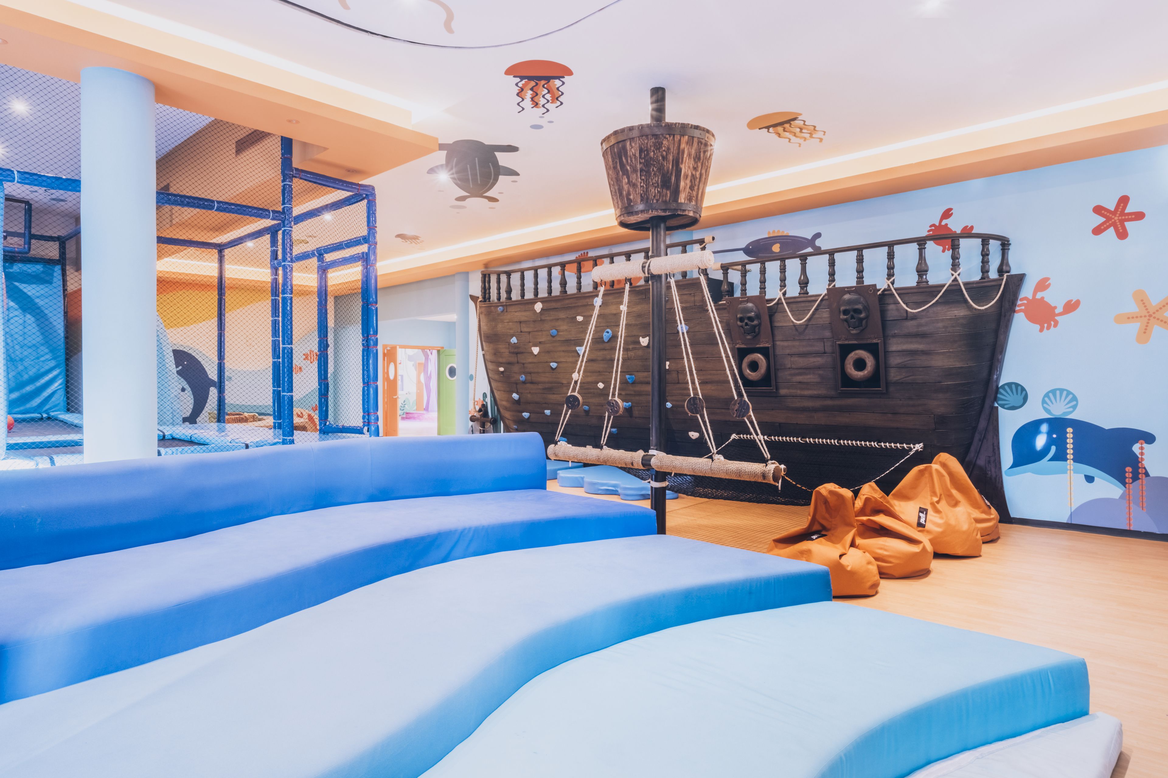 Un espace de jeux avec des marches bleues recouvertes d’un matériau amortissant, une décoration représentant un bateau en bois et une structure de jeux en forme de filet.